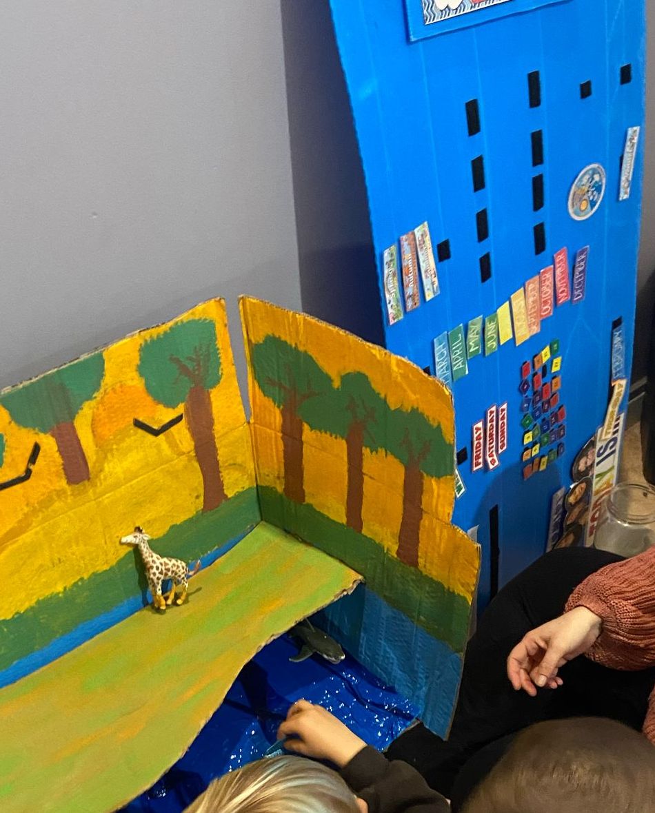A hand-painted cardboard savanna with a toy giraffe, next to a blue English class calendar with months and days
